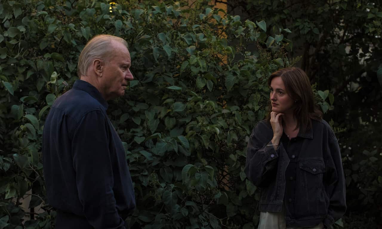 An older man in profile and a younger woman with her hand to her chin stand outdoors, facing each other against a dense backdrop of leafy green foliage.