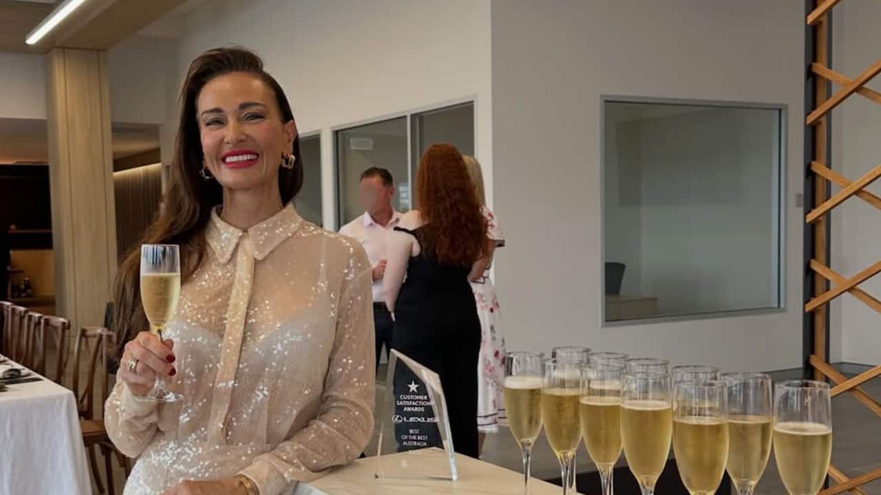 a woman in a shimmery dress smiles holding a glass of champagne in a restaurant next to a table of champagne flutes