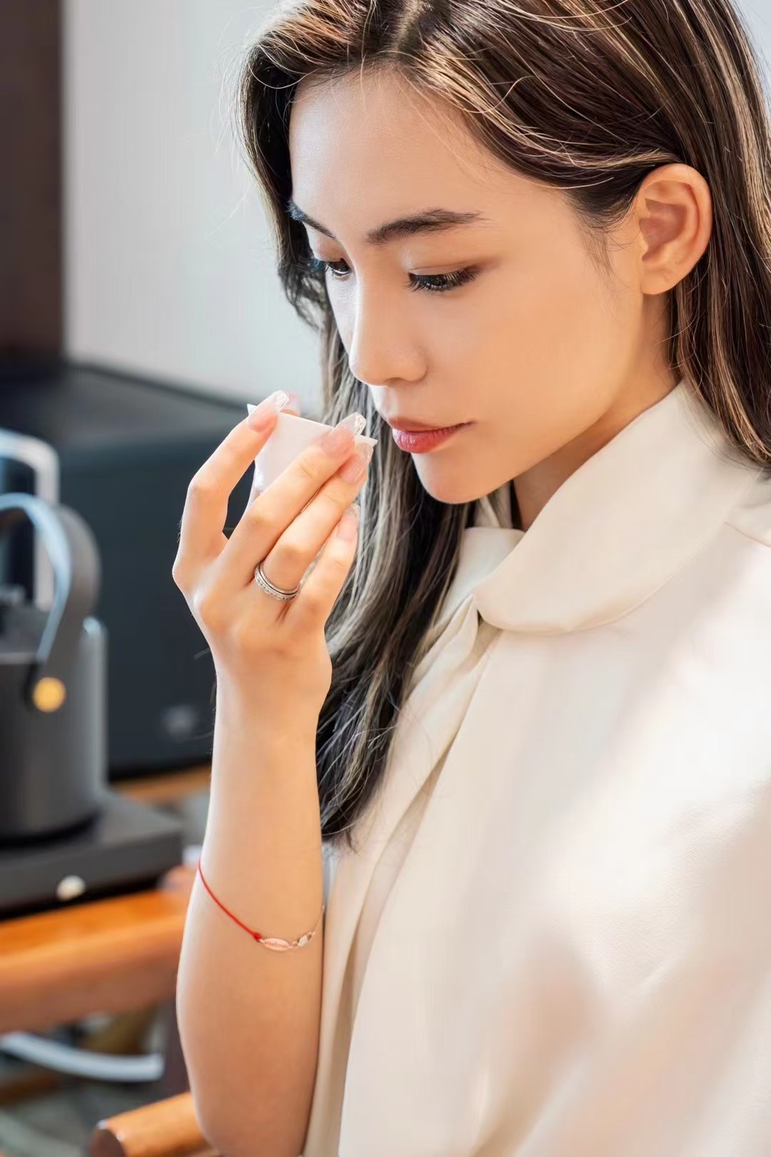 A woman wearing a white blouse sniffs a product held up to her nose in her right hand.