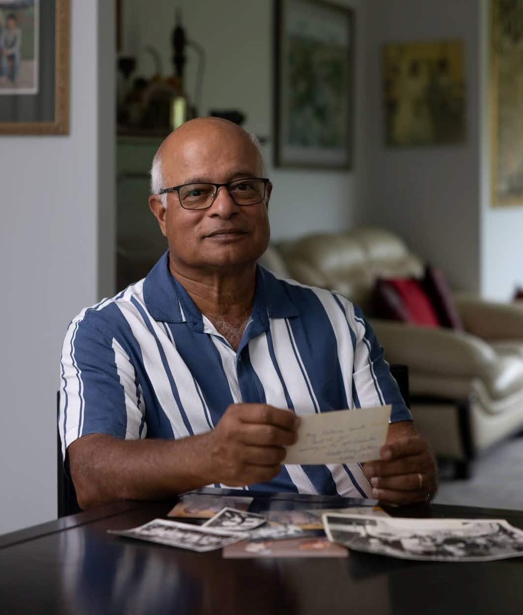 A portrait of a bald man with glasses and a blue and white striped shirt, sitting at a wooden table while holding a postcard, with old photographs scattered in front of him.