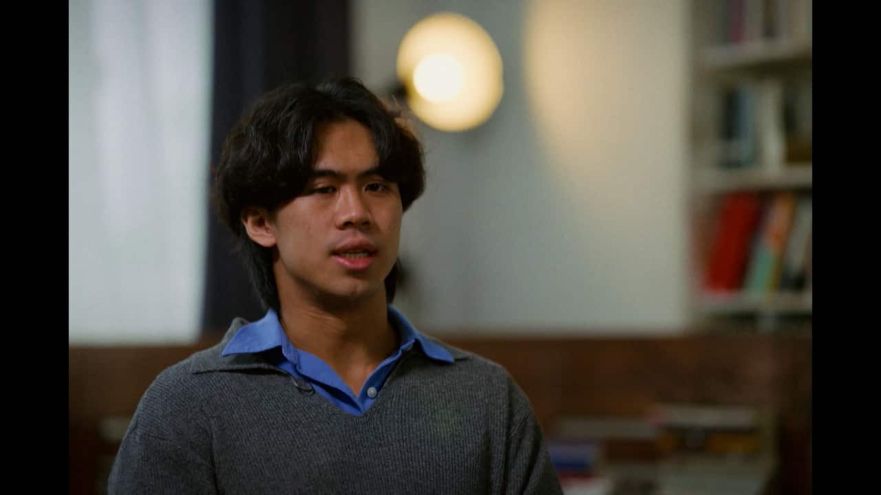 A young Asian man speaking during an interview wearing a blue collared shirt and a grey sweater