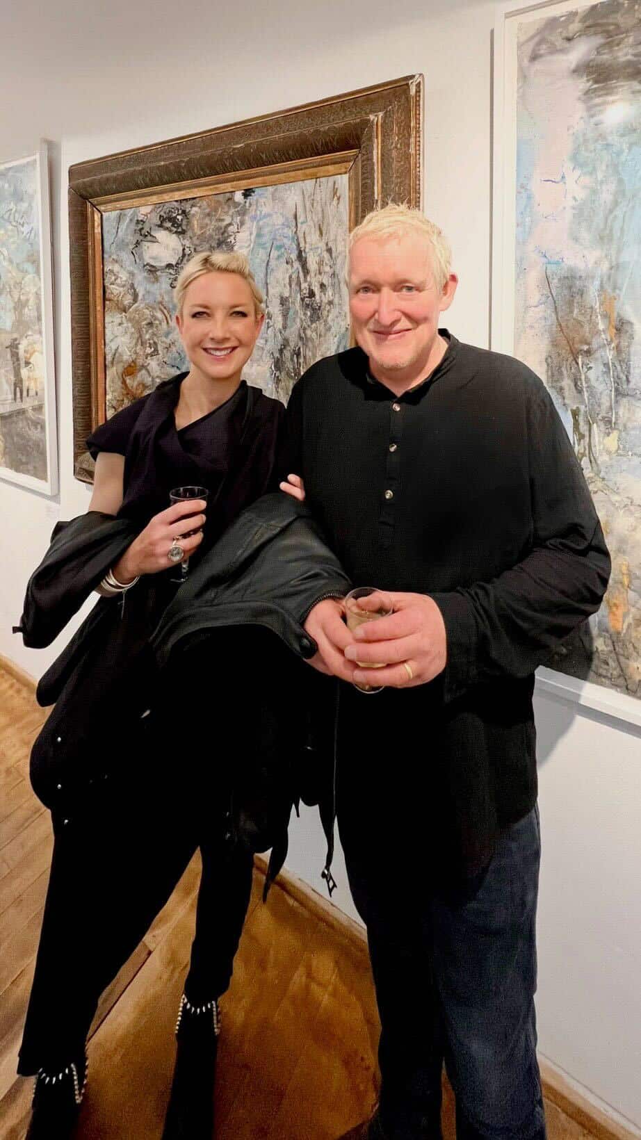 A man and a woman, both dressed in black, stand in front of paintings in an art gallery