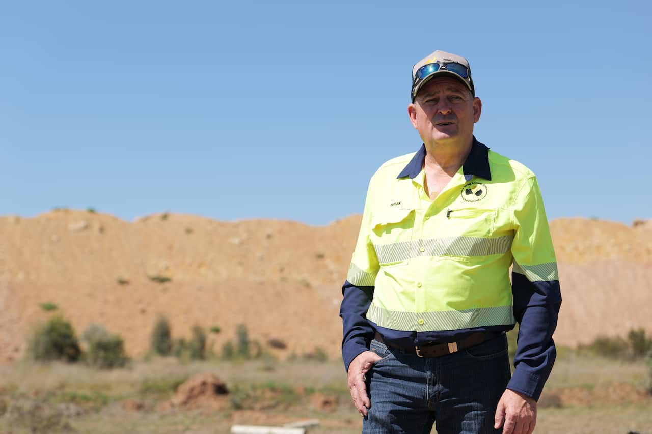 A middle-aged man stands in front of a dirt pile, wearing a yellow construction shirt, dark blue jeans, a cap and sunnies.