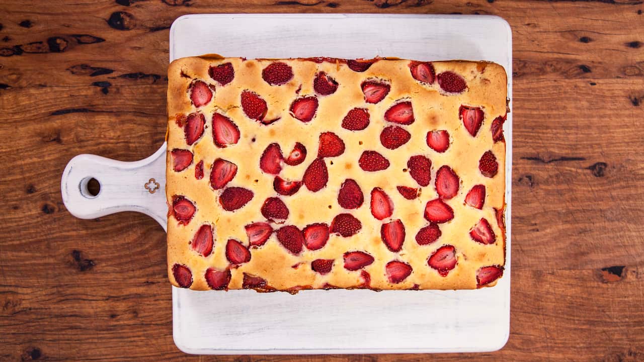 Seen from overhead, a rectangular cake studded with strawberries sits on a white paddle-shaped board. 