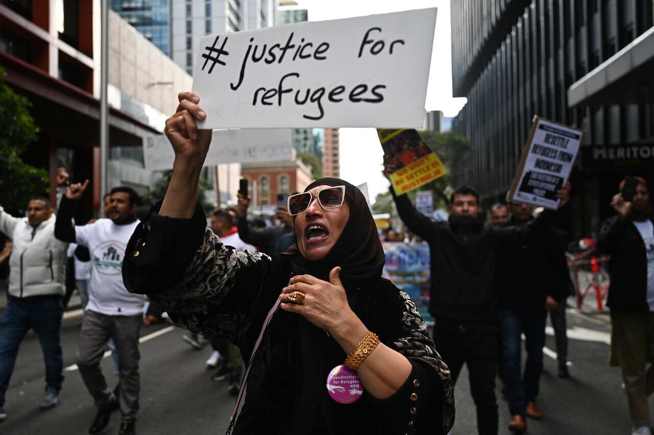 A woman in the foreground holds up a a sign that says #justice for refugees, others on the street behind her are also holding up posters.