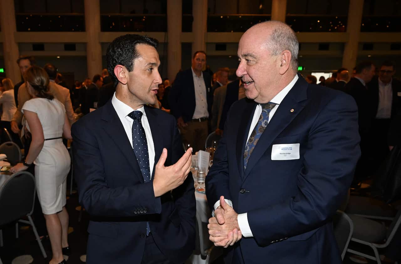 Two men in dark suits are speaking to each other in a large, indoor event space. The man on the left is gesturing with his hands while the man on the right smiles.