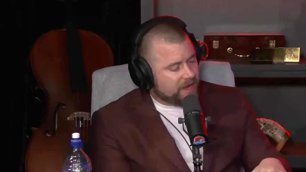 A man in a maroon blazer sitting at a desk, wearing headphones and speaking into a microphone.