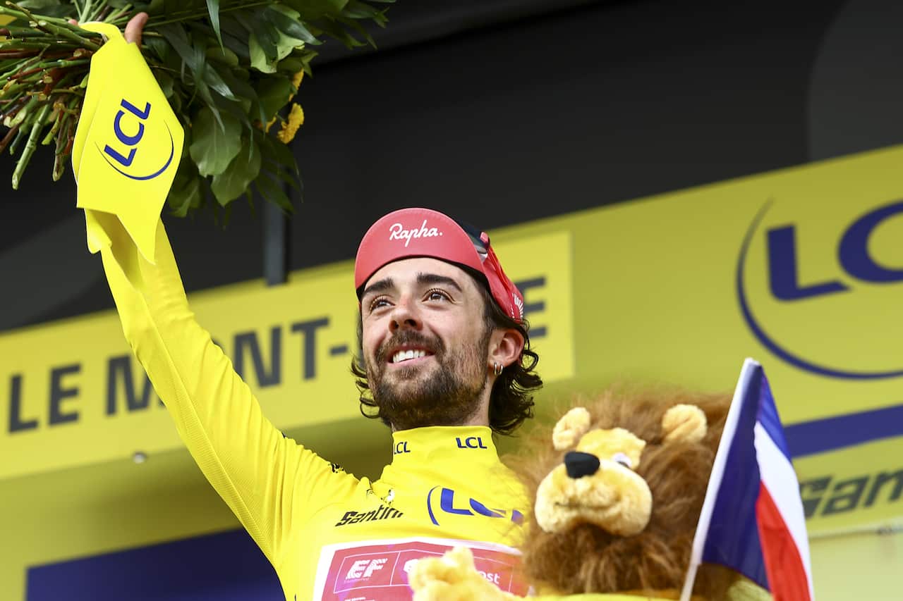 A man in a yellow jersey holds one arm up while carrying a lion plushy and a flag in the other hand.