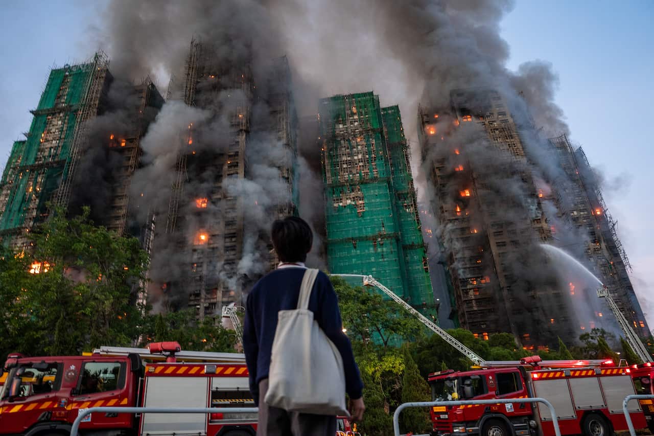 A man wearing a blue jumper and carrying a white tote bag looks on as hoses from firetrucks douse a building complex.