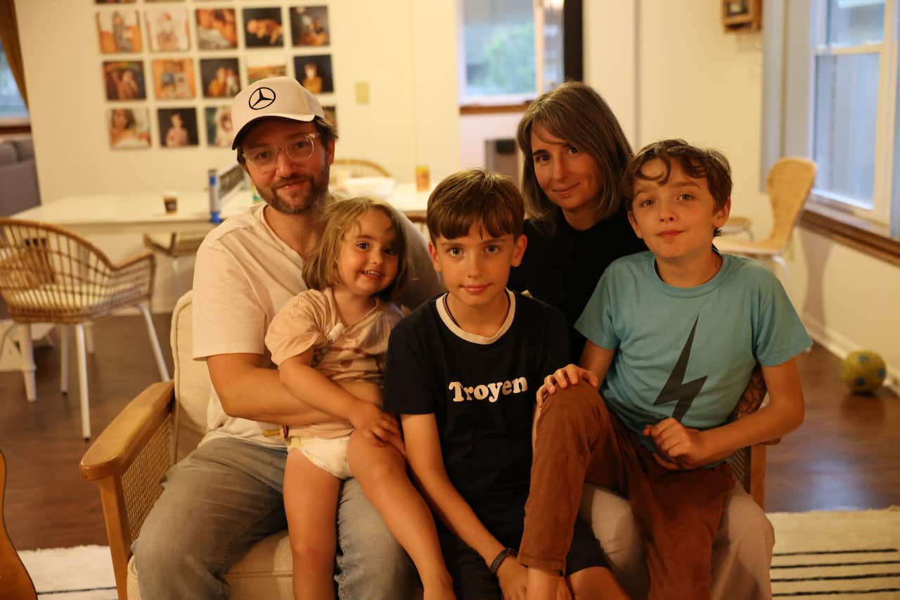 A family sits together on a couch. In the back are a father and mother. Sitting in front, from left to right, are a toddler-aged girl and two young boys. 