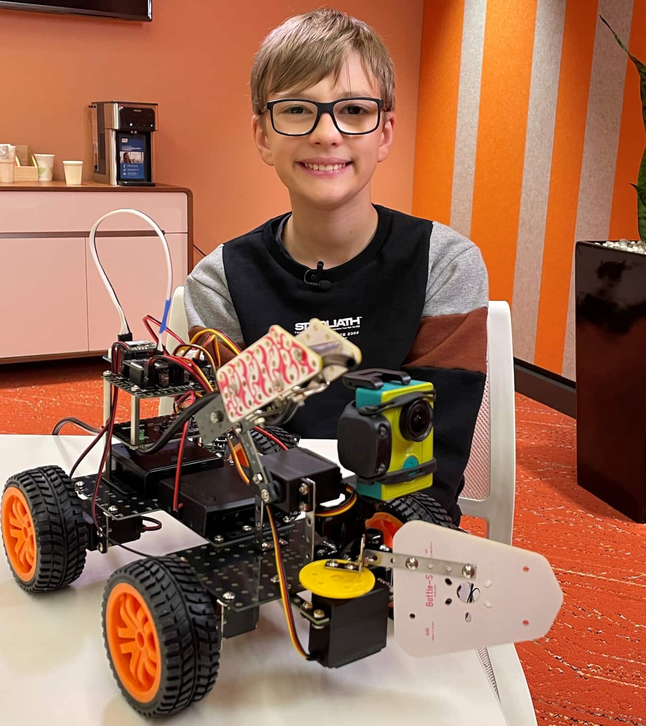 A boy smiling behind a robot with four wheels on a table 