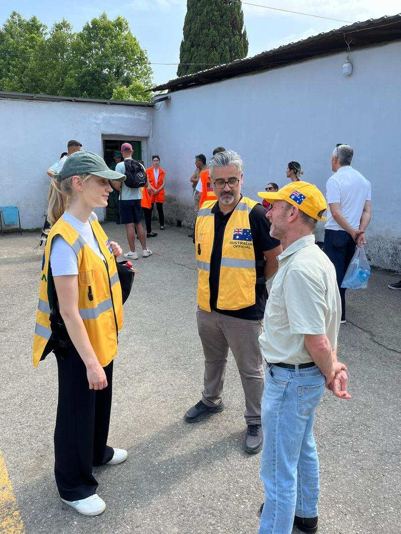 Two men and a woman are standing in a circle and talking. There are a few people standing behind them.