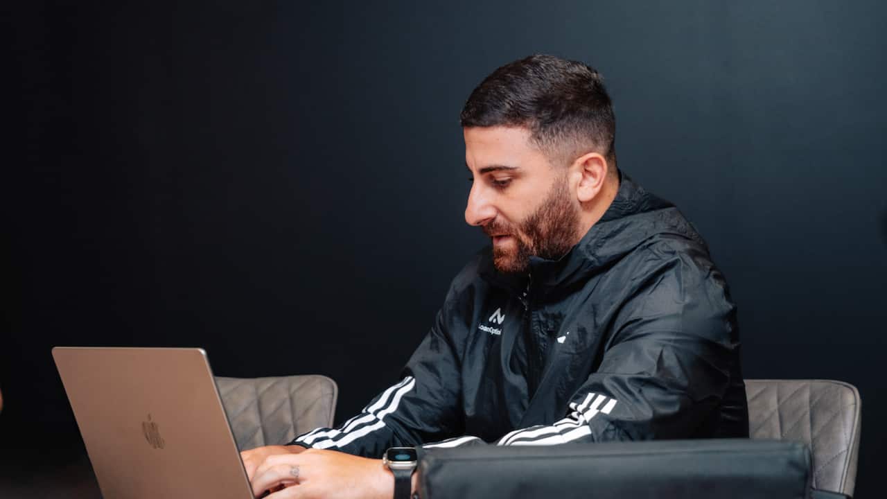 a man in his thirties with a brown hair and beard working on a laptop in a black office