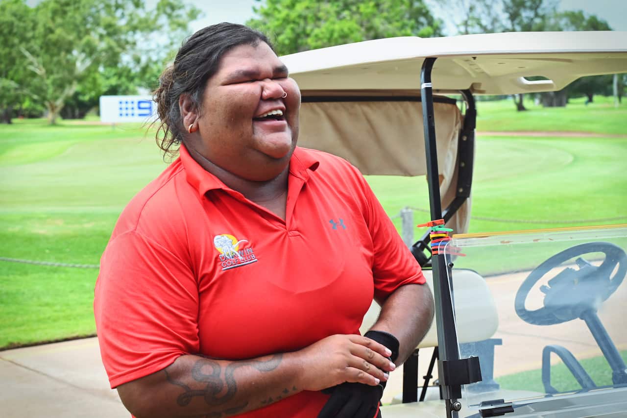 A woman wearing a red shirt stands next to a golf cart.