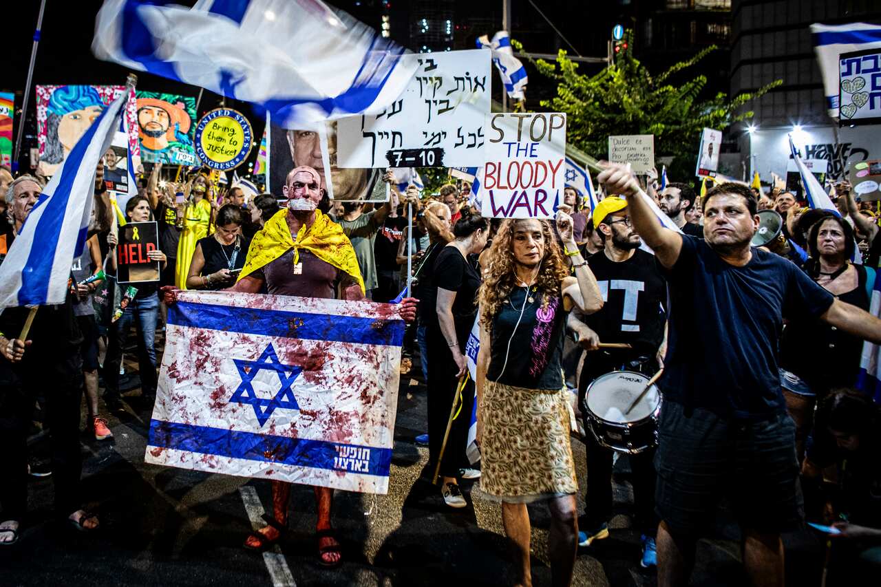 Protesters marching through the street, many holding signs. One person is holding an Israeli flag stained with fake blood.