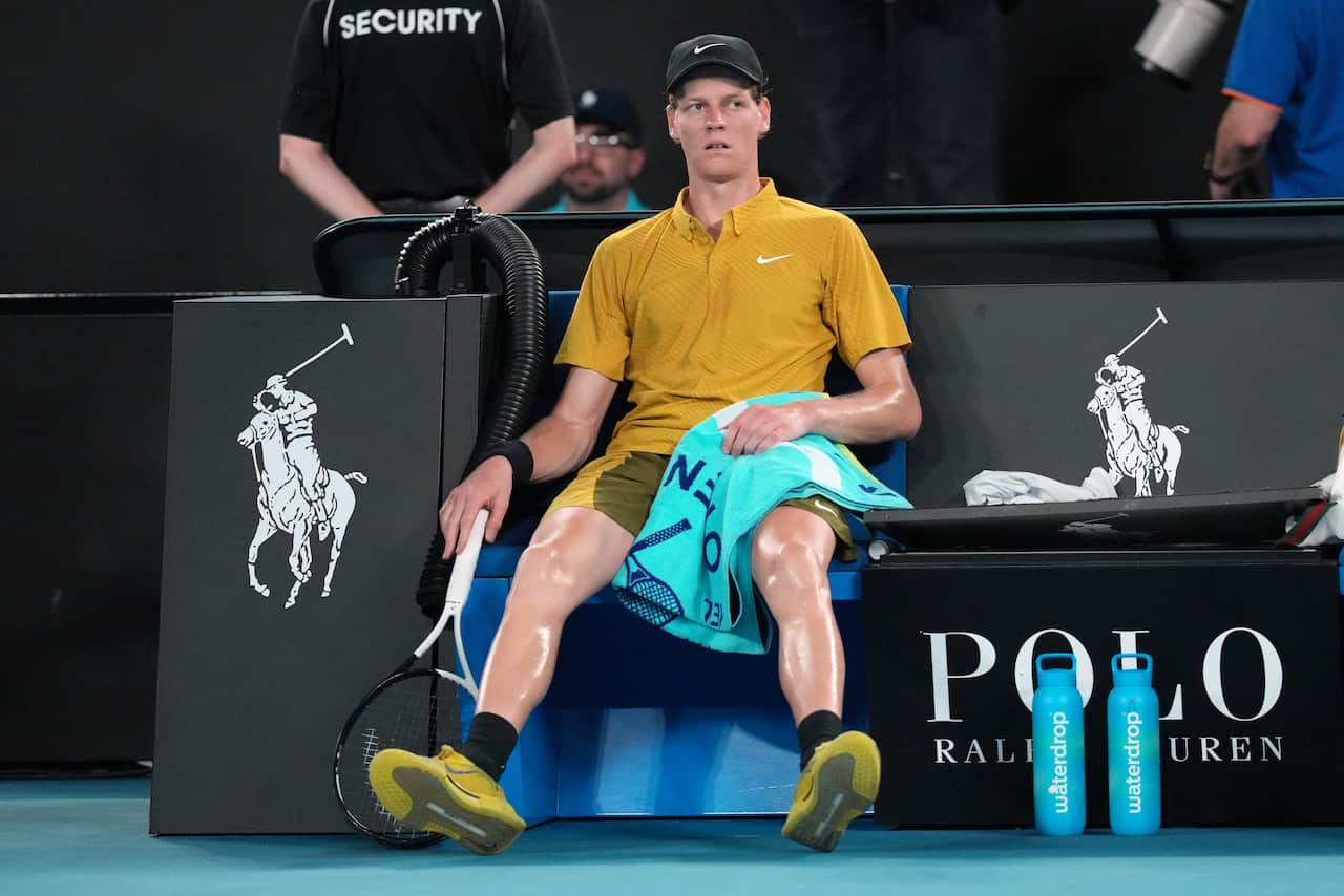 A tennis player sits on the bench, racket on the ground and towel in his lap, recovering from heat stress.