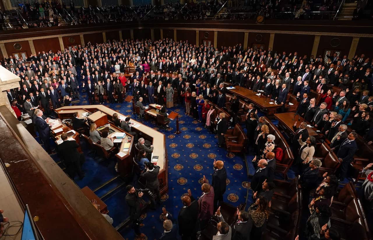 A room of people wearing suits stand as they are sworn in as members of the US House of Representatives.