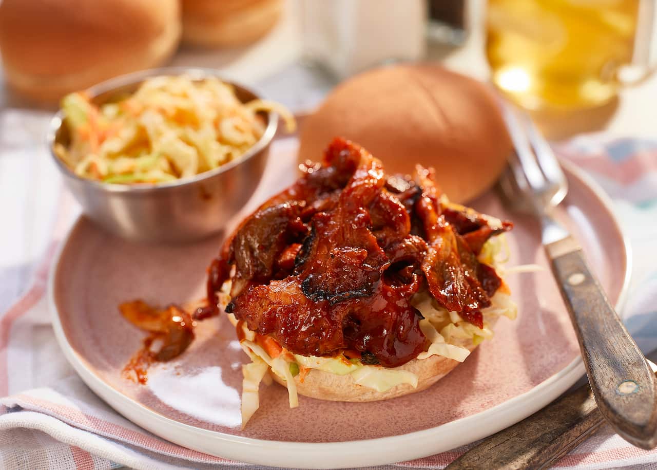 A burger bottom is topped with a little coleslaw and a mound of juicy, sticky mushroom pieces. The burger top sits alongside. More buns and coleslaw can be seen in the background.