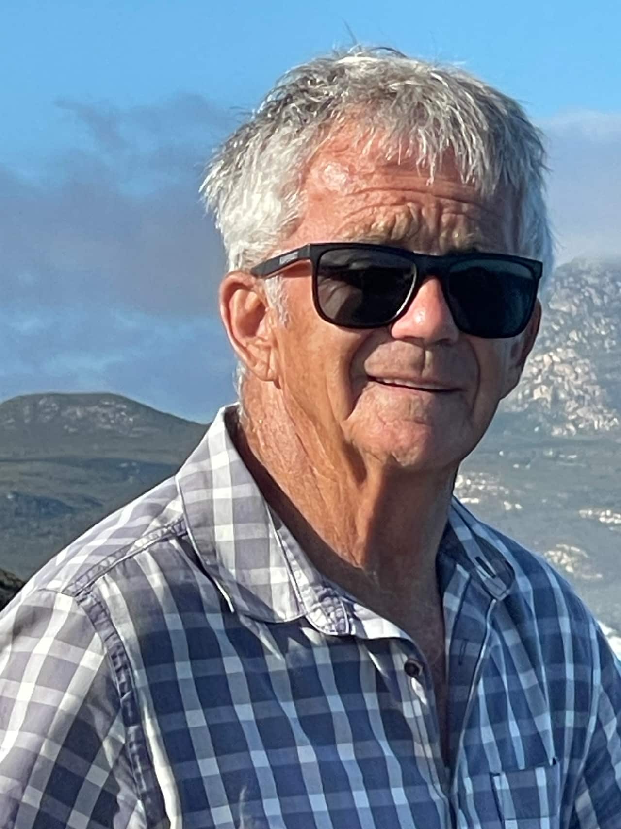Close up of a man with short grey hair and wearing dark sunglasses and a grey and white check shirt standing outside with hills in the background