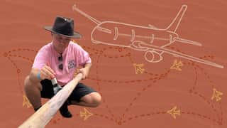 A man wearing a pink T-shirt and a brown hat is painting a yidaki. There are sketches of some small and a big airplane in the background.