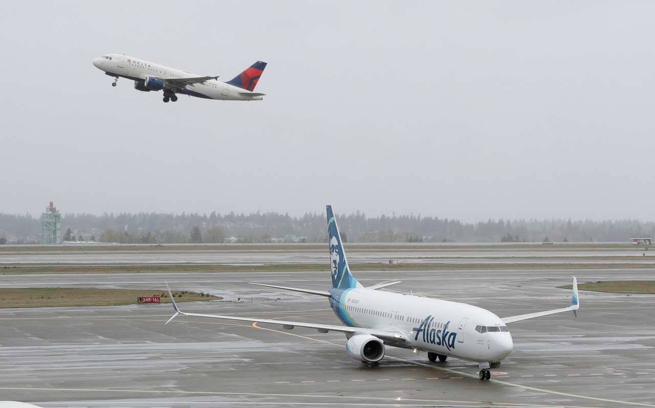 Grounded white and blue airplane sits on tarmac at airport. Skyline visible in background. A separate plane flies off in the distance.