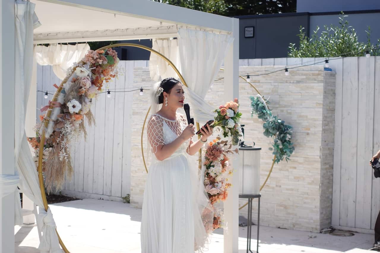 A woman wearing a wedding dress standing outside and speaking into a microphone.