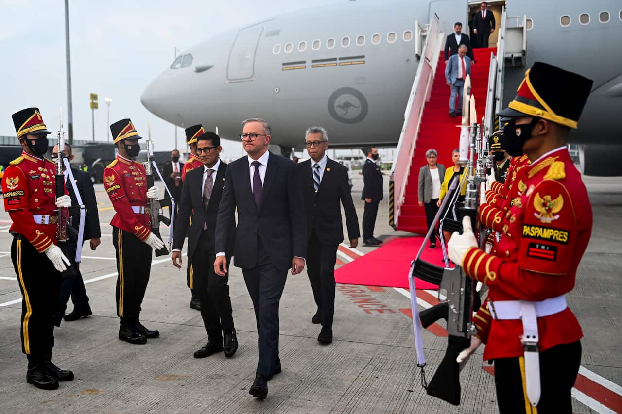 A group of men in suits walking on the tarmac from a plane. Soldiers in red ceremonial uniform form a guard of honour