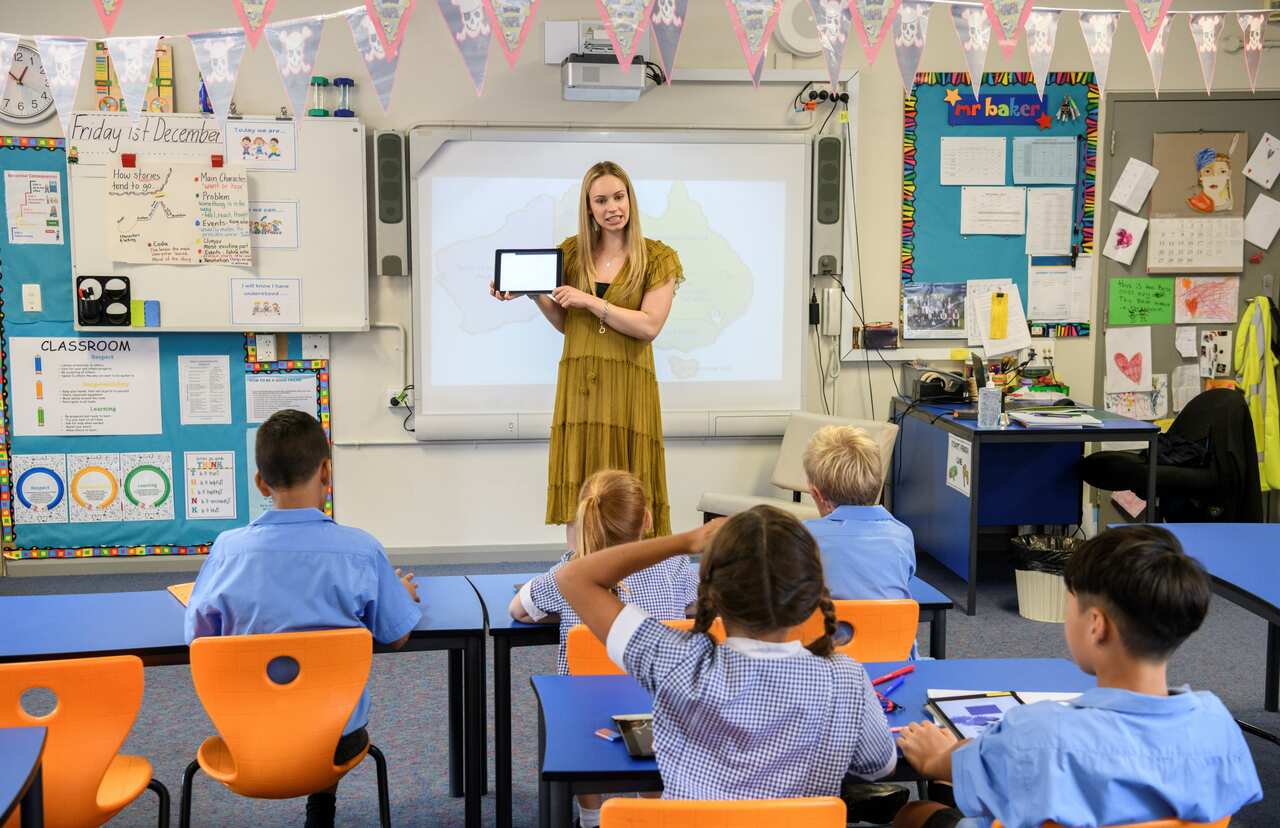 Children sitting at desks listening to teacher holding digital tablet