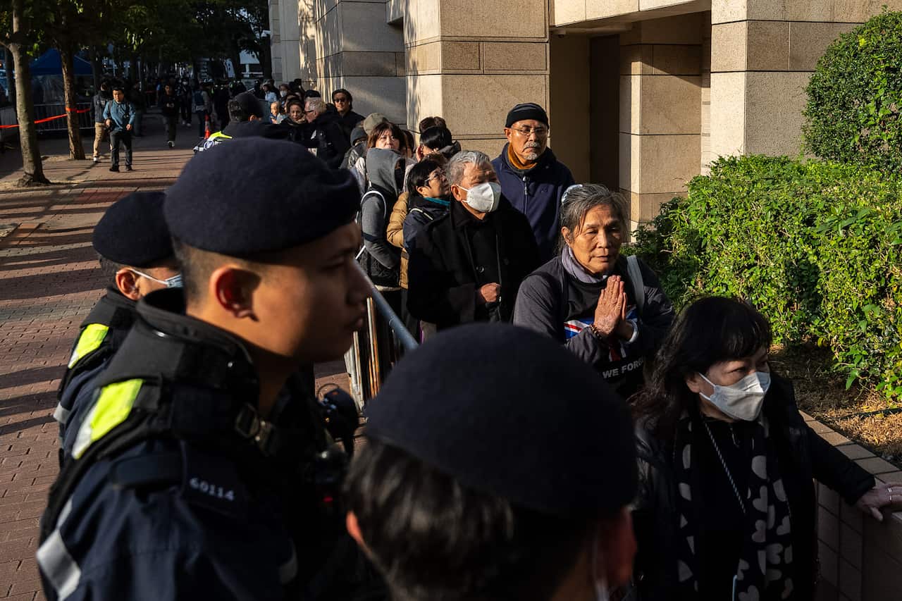 A line of people, some wearing face masks, waits behind a metal barricade outside a building while being monitored by police officers in blue uniforms.