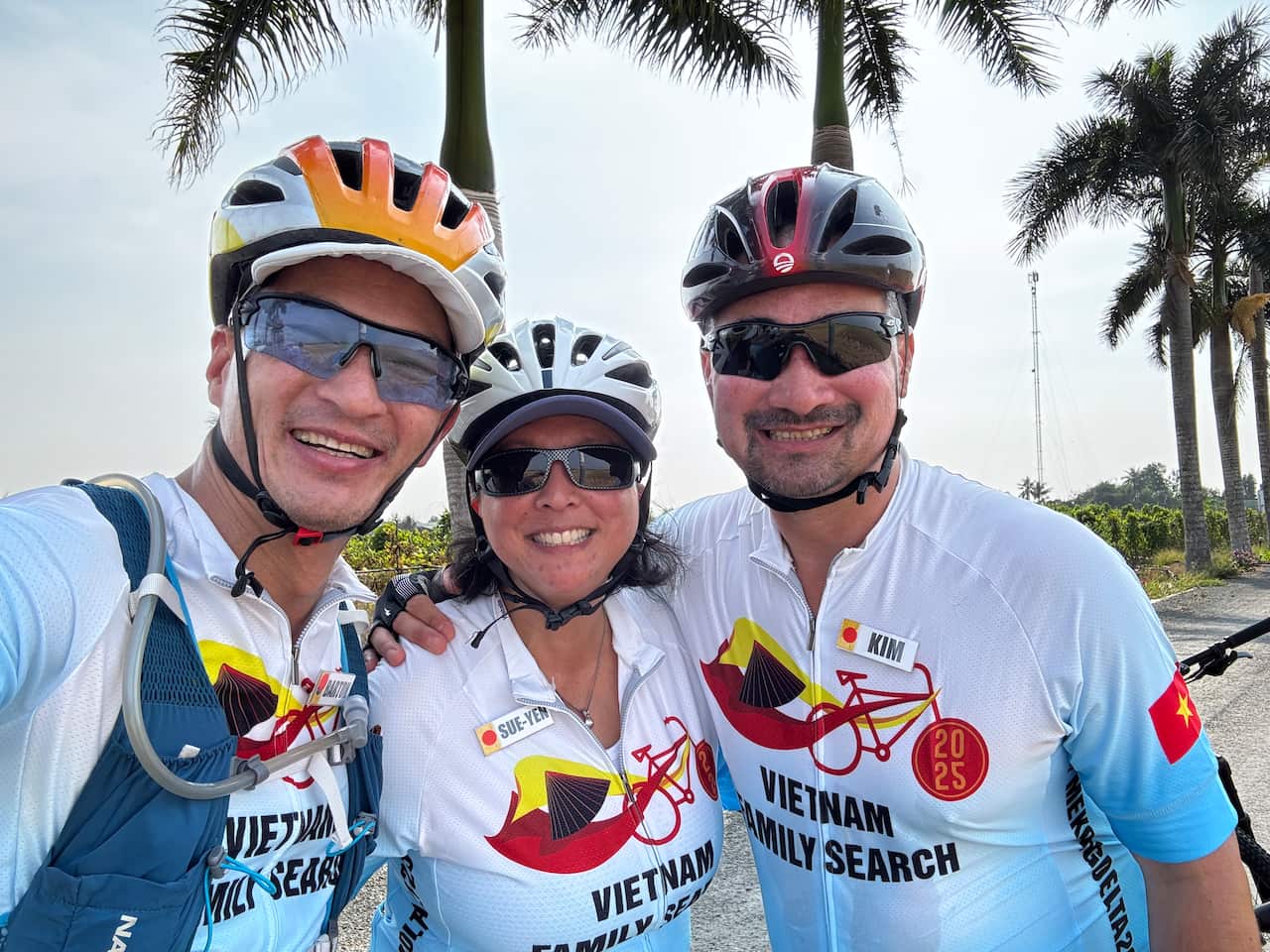 A middle-aged man, a middle-aged woman in the centre and another middle-aged man, all of Asian descent, wearing bike riding glasses and blue and white shirts that read 'Vietnam Family Search', smiling at the camera, with palm trees in the background.