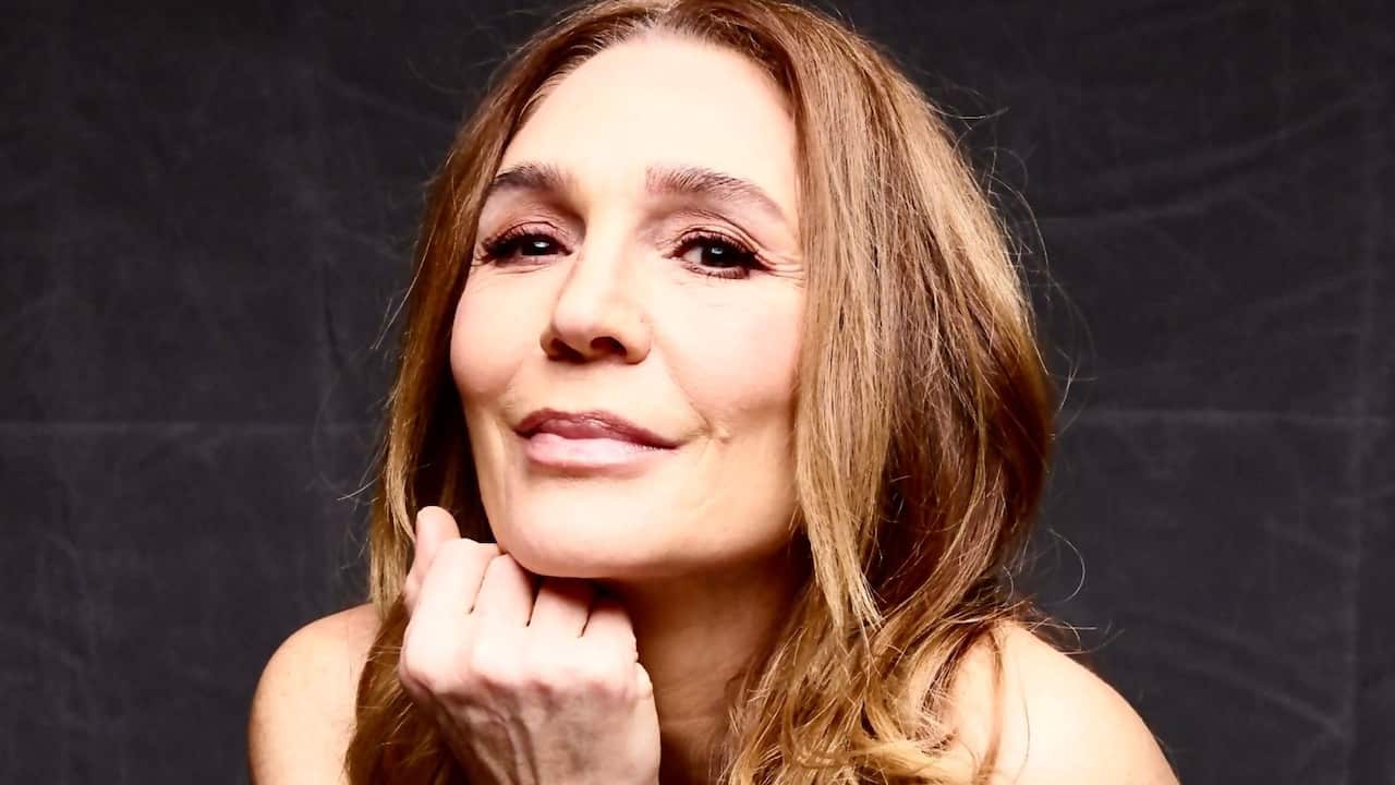 a middle aged woman with long light brown hair with her chin on her fist, smiling at the camera in front of a black studio background