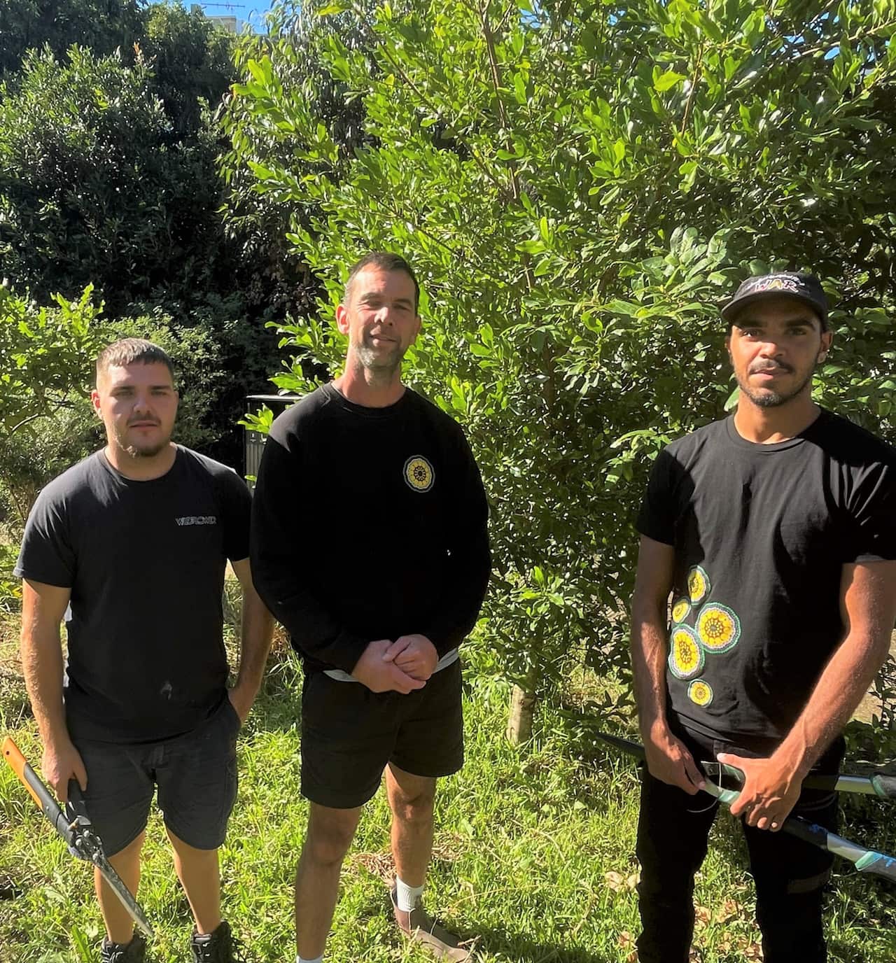 Three men stand in a park in front of big trees.