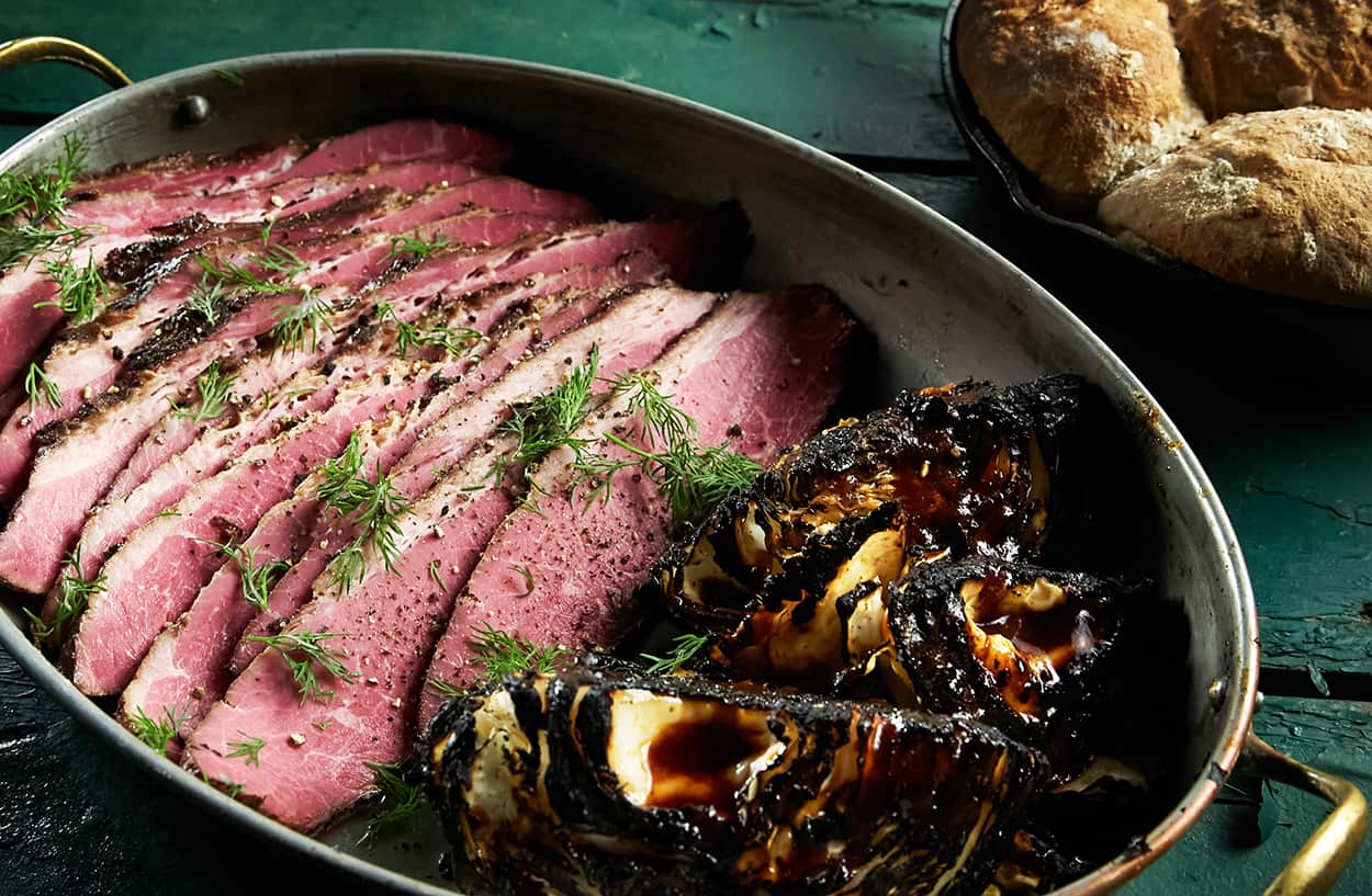 An oval metal tray, sitting on a green-painted wooden surface, holds sliced corn beef and charred cabbage wedges. 