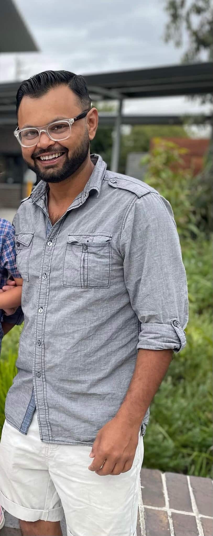 A man wearing glasses, a grey long-sleeved shirt and white shorts smiles for a photo. There is a garden behind him.