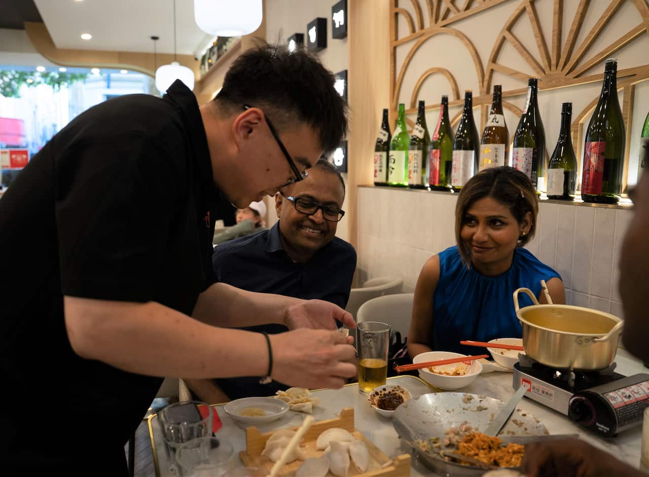 A man in a black t-shirt leans over a table showing customers how to make dumplings.