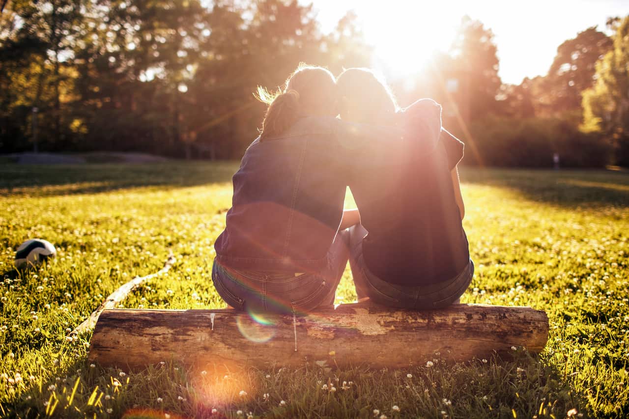 Rear view of affectionate sisters sitting on log at park during sunny day