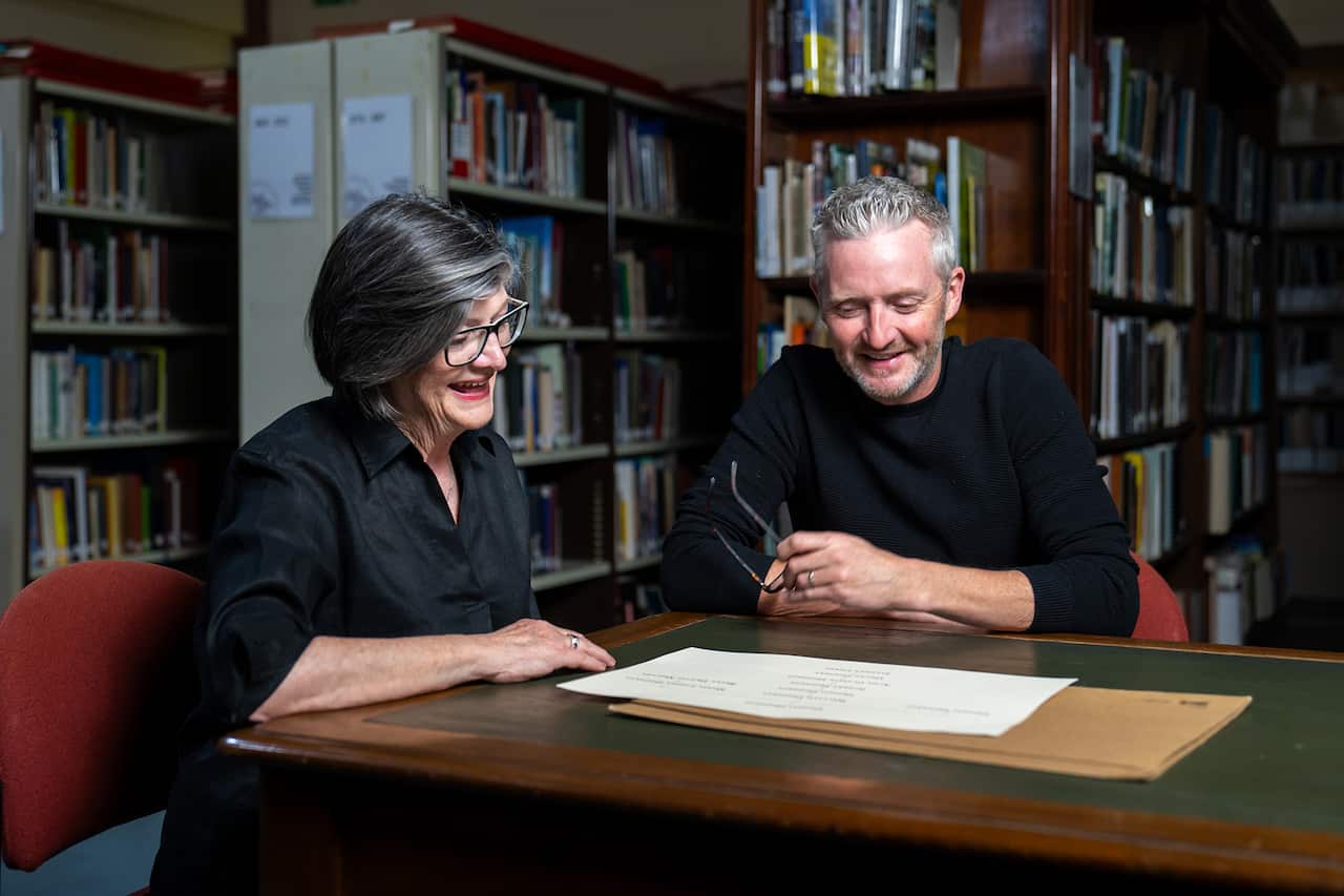 A man and a woman sit at a table in a library. They are looking at a dicusment that sits in front of them. 