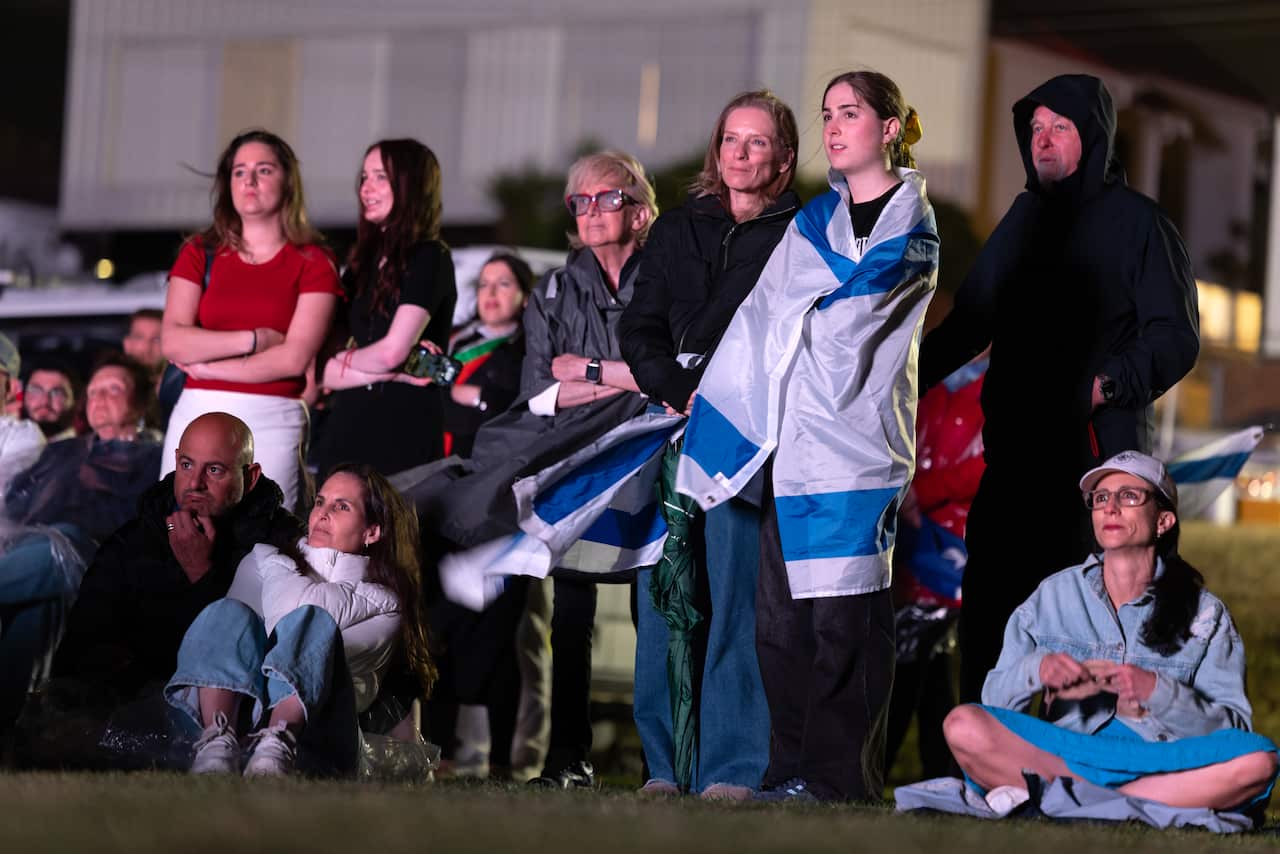 A diverse group of people, some seated on the grass and others standing, are gathered outdoors at night, intently focused on something out of frame. A young woman in the centre is wrapped in a large white and blue Israeli flag.