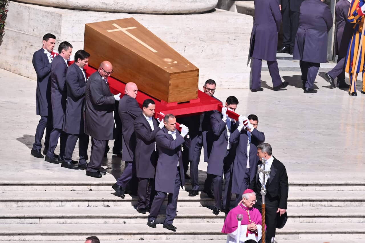 A group of men wearing dark coloured formal suits carry a wooden coffin down a set of stairs.