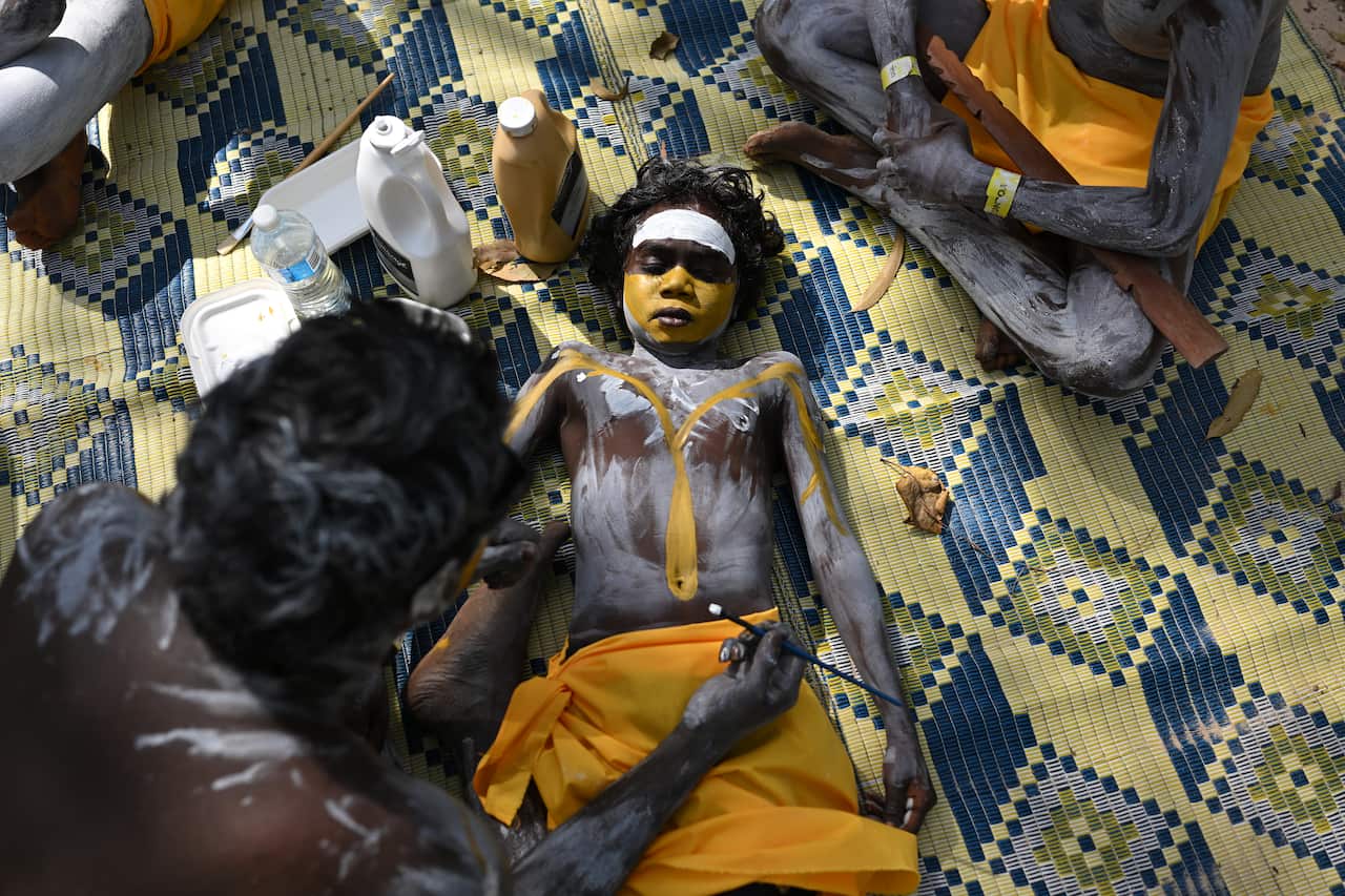 A young boy lies on the ground while getting yellow and white paint applied to his body.