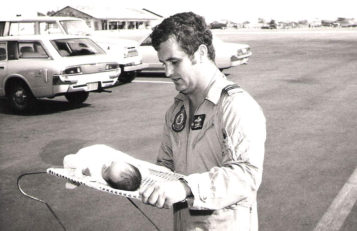 A pilot in a jumpsuit carrying a baby in a bassinet in an outdoor carpark