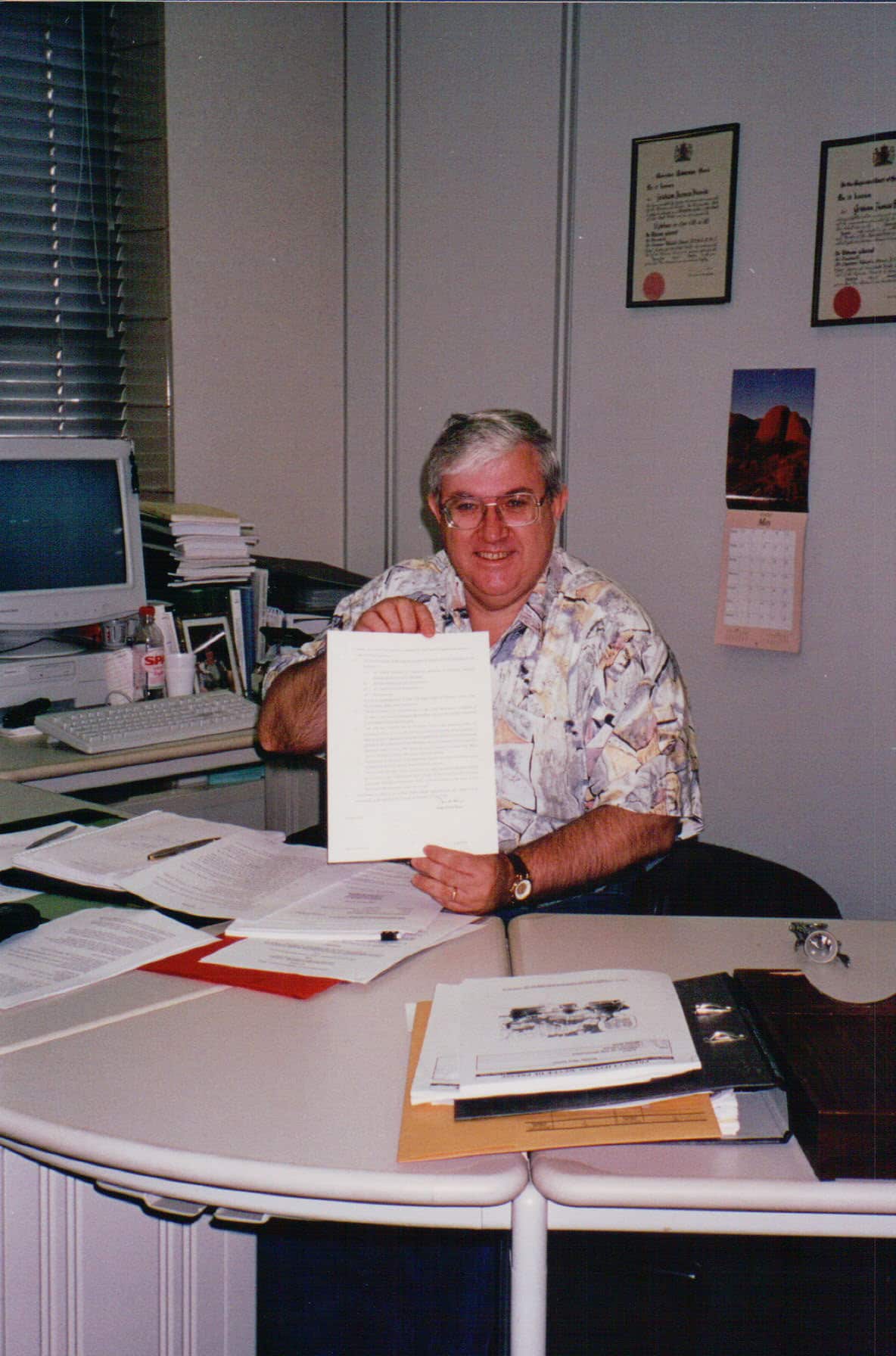 Older, largish man with white hair, wearing a casual patterned shirt, at a desk with papers on it, holding up a piece of paper. 