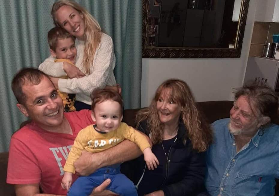 A man is sitting on a couch holding a child and there is a woman and a man sitting next to them. A woman holding a child is behind them.