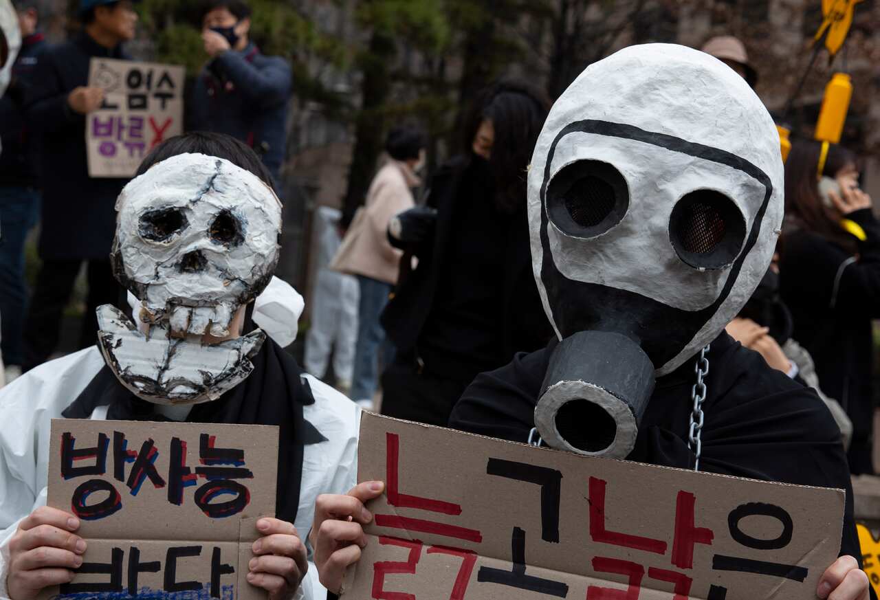 A person in a skull mask and another person in a handmade gas mask stand side by side holding signs written in Korean.