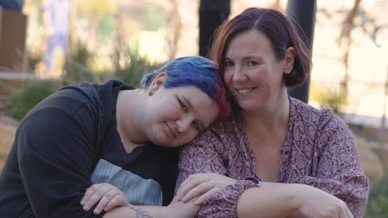 A person with blue and pink hair, sitting with their head resting on the shoulder of a woman with short hair.