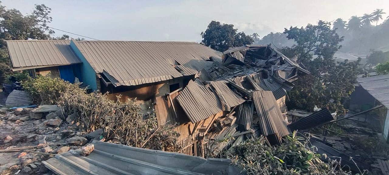 Damaged houses with tin roofs are surrounded by tall trees.