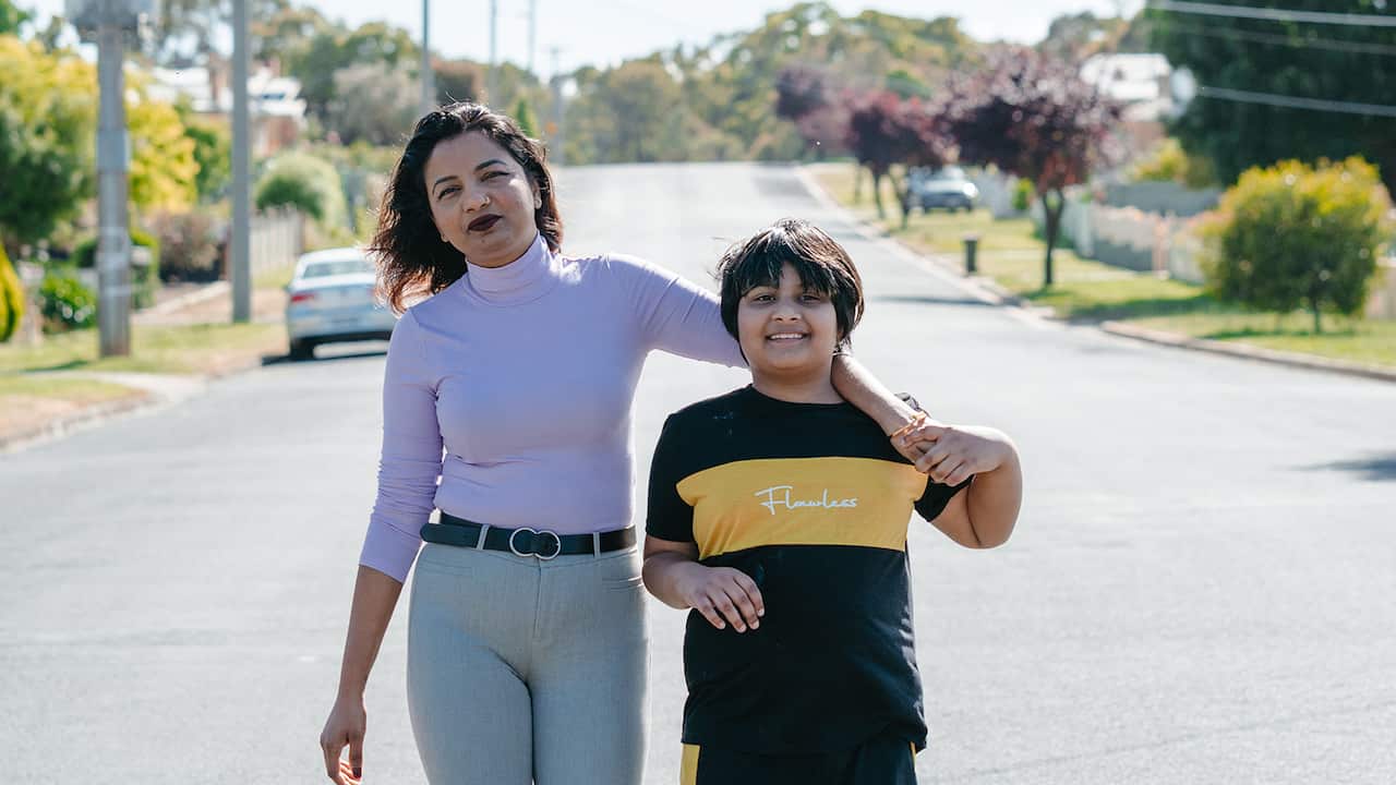 A woman stands with her arm around a boy at the end of a suburban street. They are both smiling. 