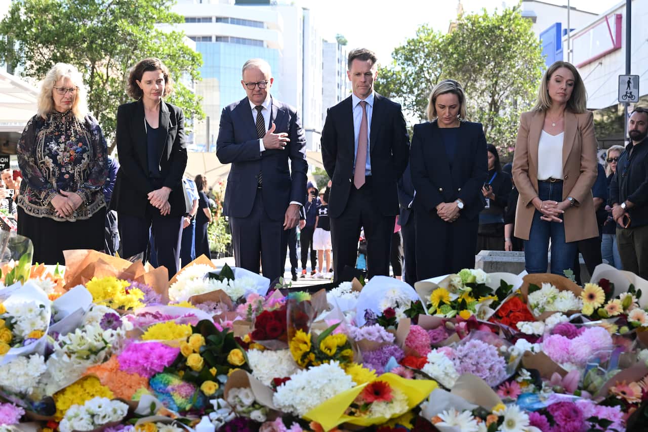 A group of people, including Prime Minister Anthony Albanese and NSW Premier Chris Minns, stand in front of a large floral tribute.