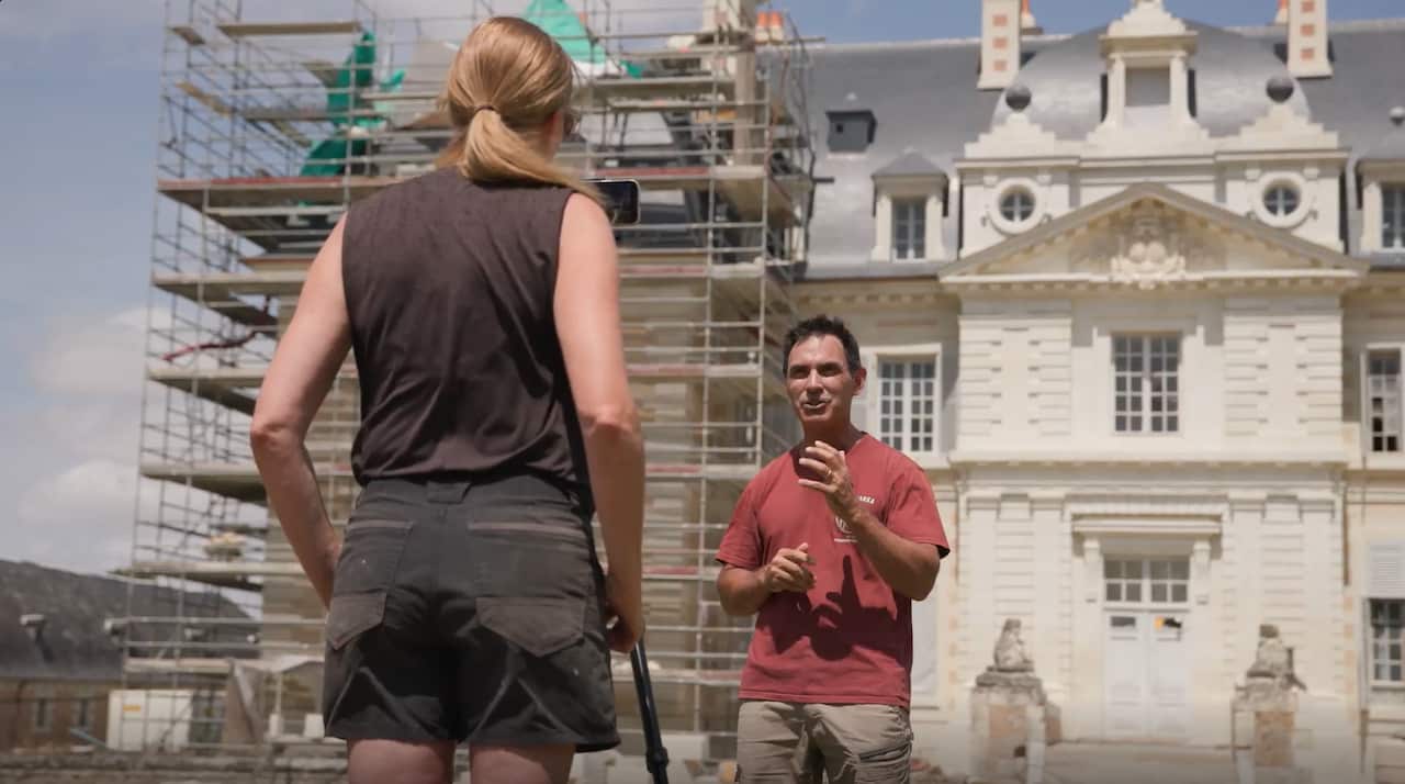 A man and a woman stand in front of a grand house, half of which has scaffolding on it. The woman has her back to the viewer and is looking at a phone on a tripod. The man is facing her, speaking and gesticulating.