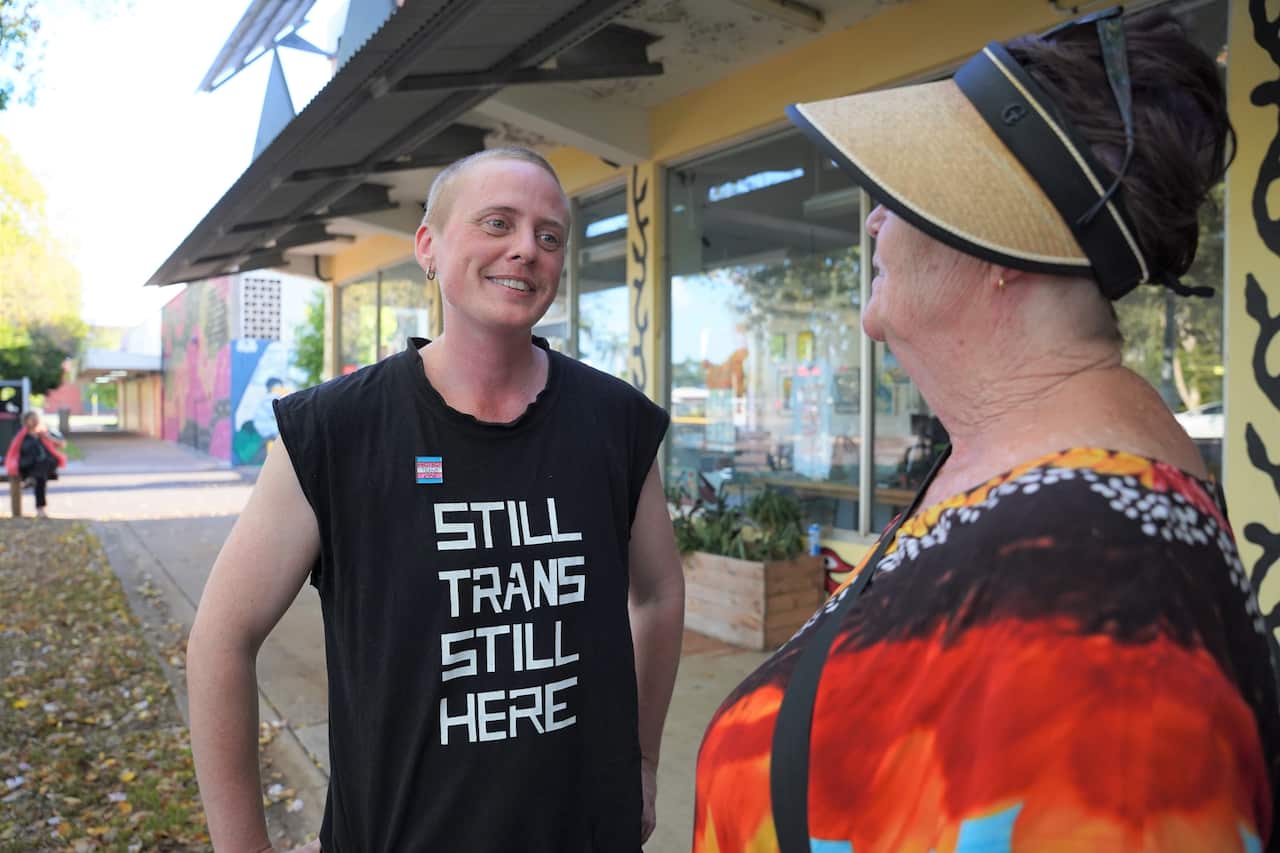 [JVS] A person wearing a black sleeveless T-shirt with the words "still trans, still here" written in white in all-caps font.